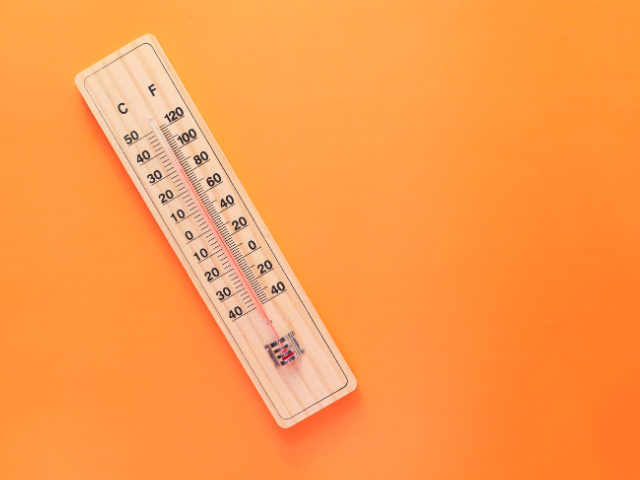 A wooden thermometer showing high temperatures in both Celsius and Fahrenheit against a bright orange background.