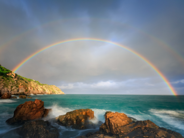 A vivid rainbow arcs over the ocean with rocky cliffs in the foreground, under a cloudy sky.